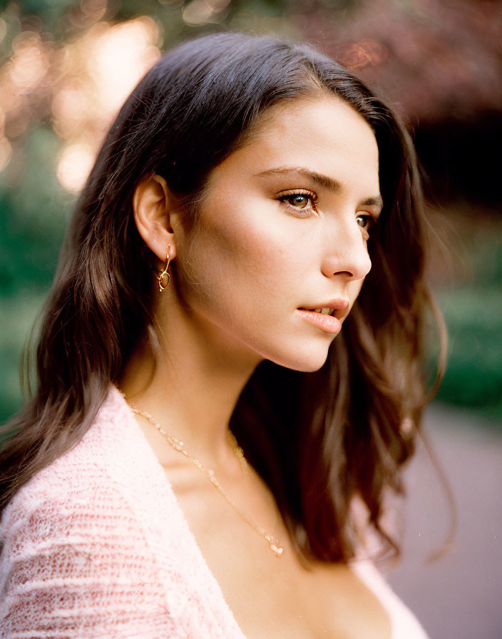 Portrait of woman with long dark hair in pink sweater, gazing thoughtfully in garden setting