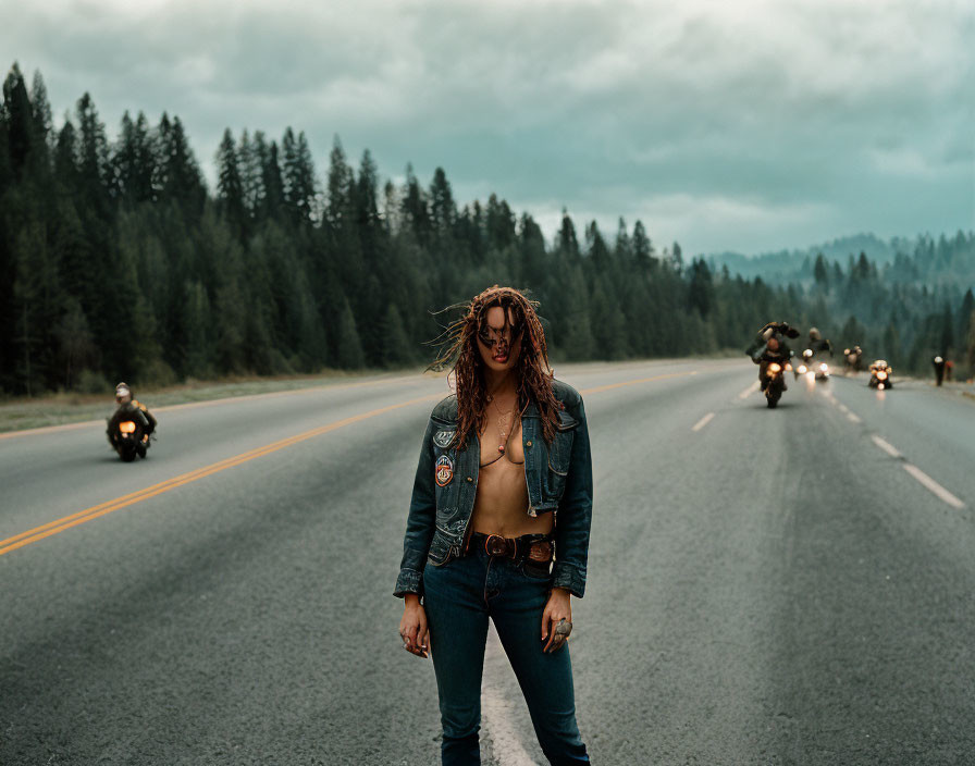 Long-haired person in denim with patches on road, motorcyclists in forested background