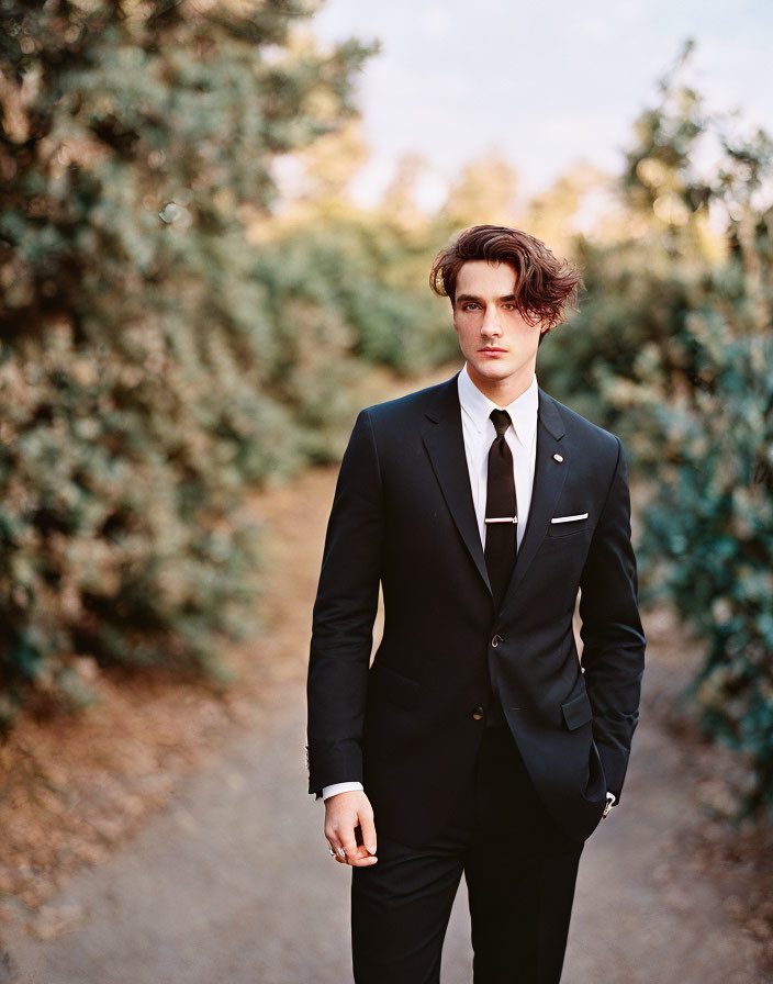 Wavy-haired man in black suit on path with trees