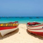 Wooden boats with red flowers on sandy beach, turquoise sea, sailing boats, daytime moon.