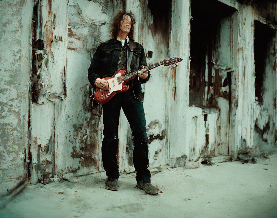 Long-haired man playing electric guitar against weathered wall.