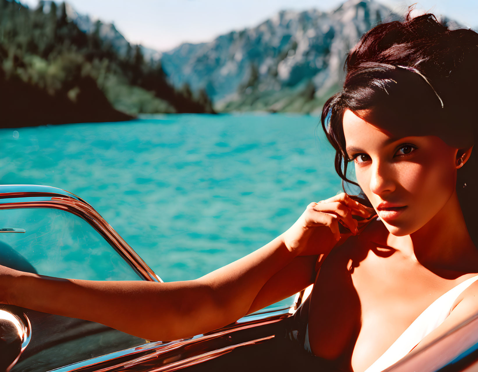 Woman posing in vintage convertible by turquoise lake with mountains in background.
