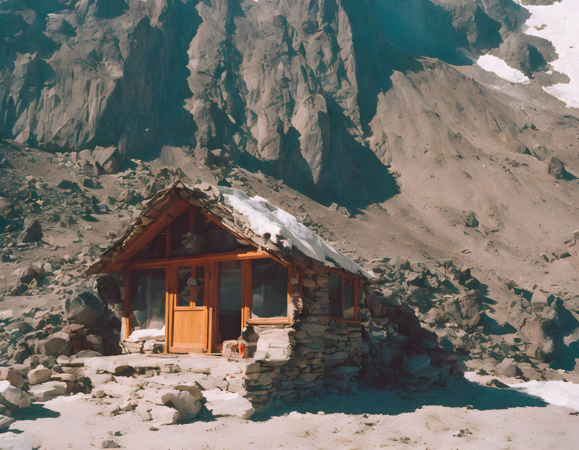 Stone cabin with wooden door and windows at mountain base in snow.