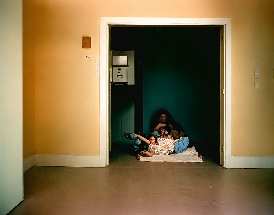 Three people cuddled on a blanket in a dark room with bright orange wall.