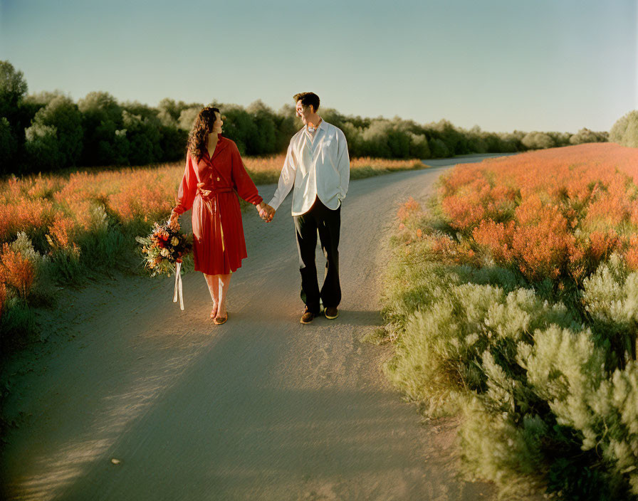 Couple walking hand in hand on dirt road with orange-blossomed bushes and clear blue sky