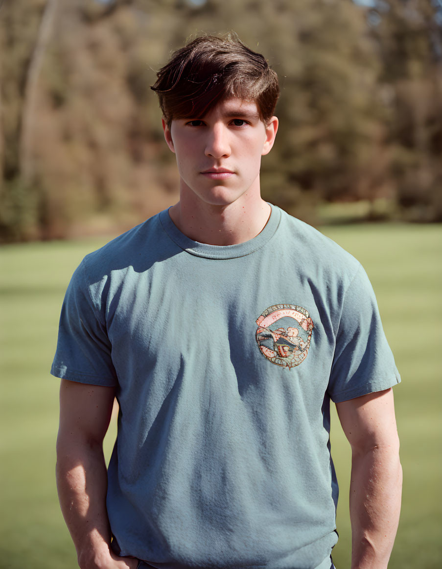 Young man with dark hair in blue t-shirt outdoors with trees and grass