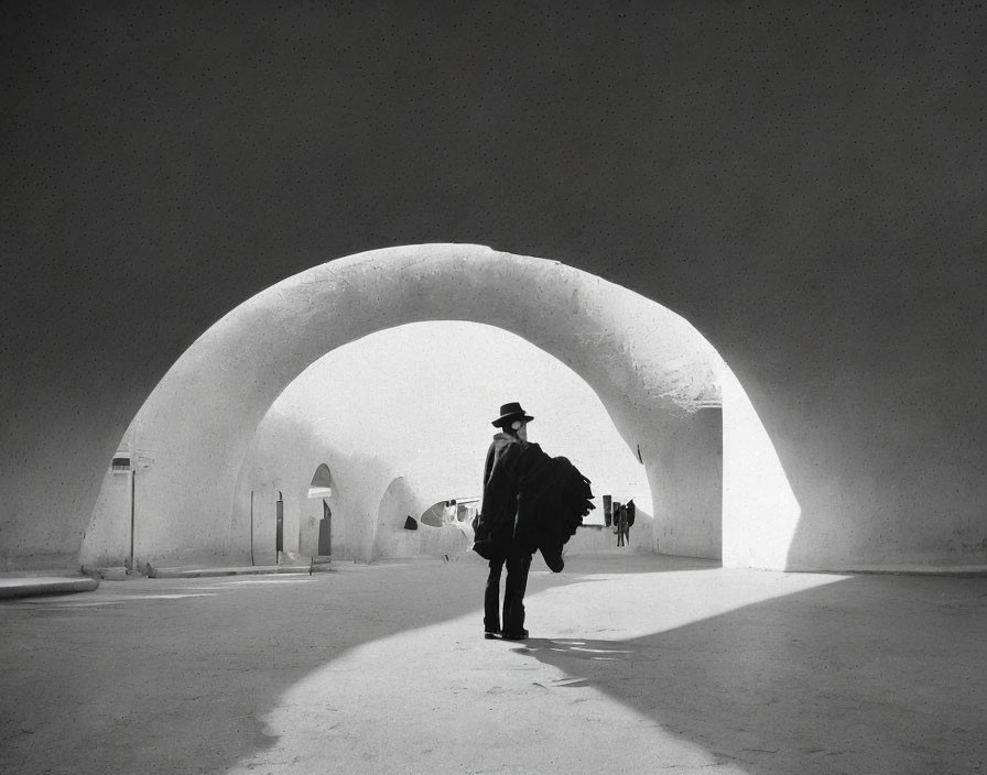 Person in dark attire casting shadow under large archway in monochrome setting