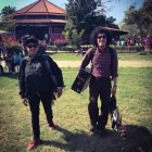 Two individuals with speaker heads and electric guitars in stylish outfits at a park with a futuristic building.