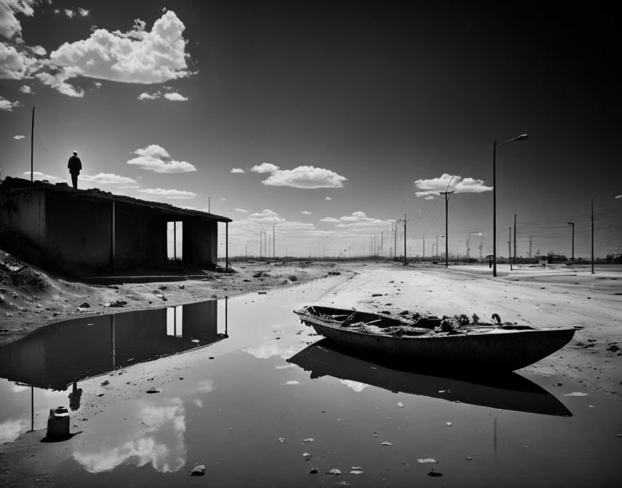 Monochrome image: person by stranded boat on dry landscape with reflecting water & vast sky.