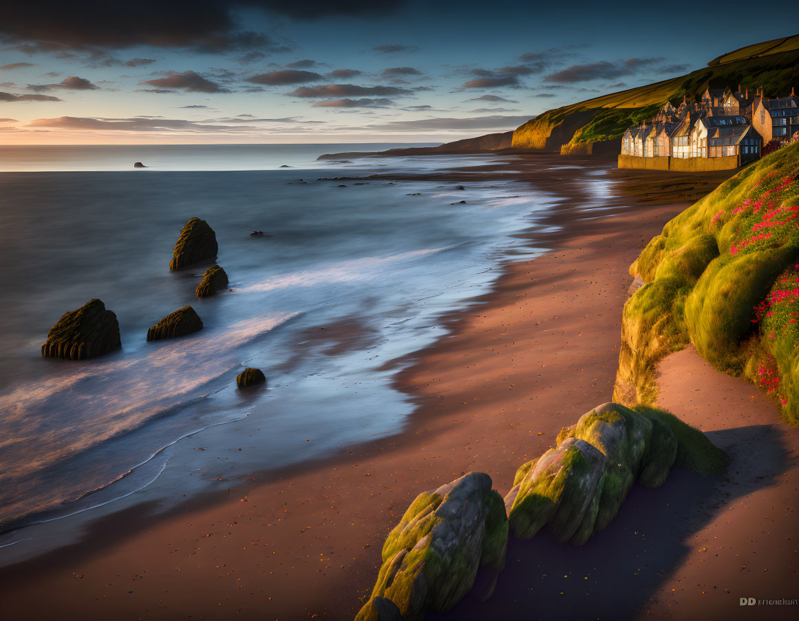 Tranquil beach scene at sunset with rocky outcrops and houses nestled among green cliffs