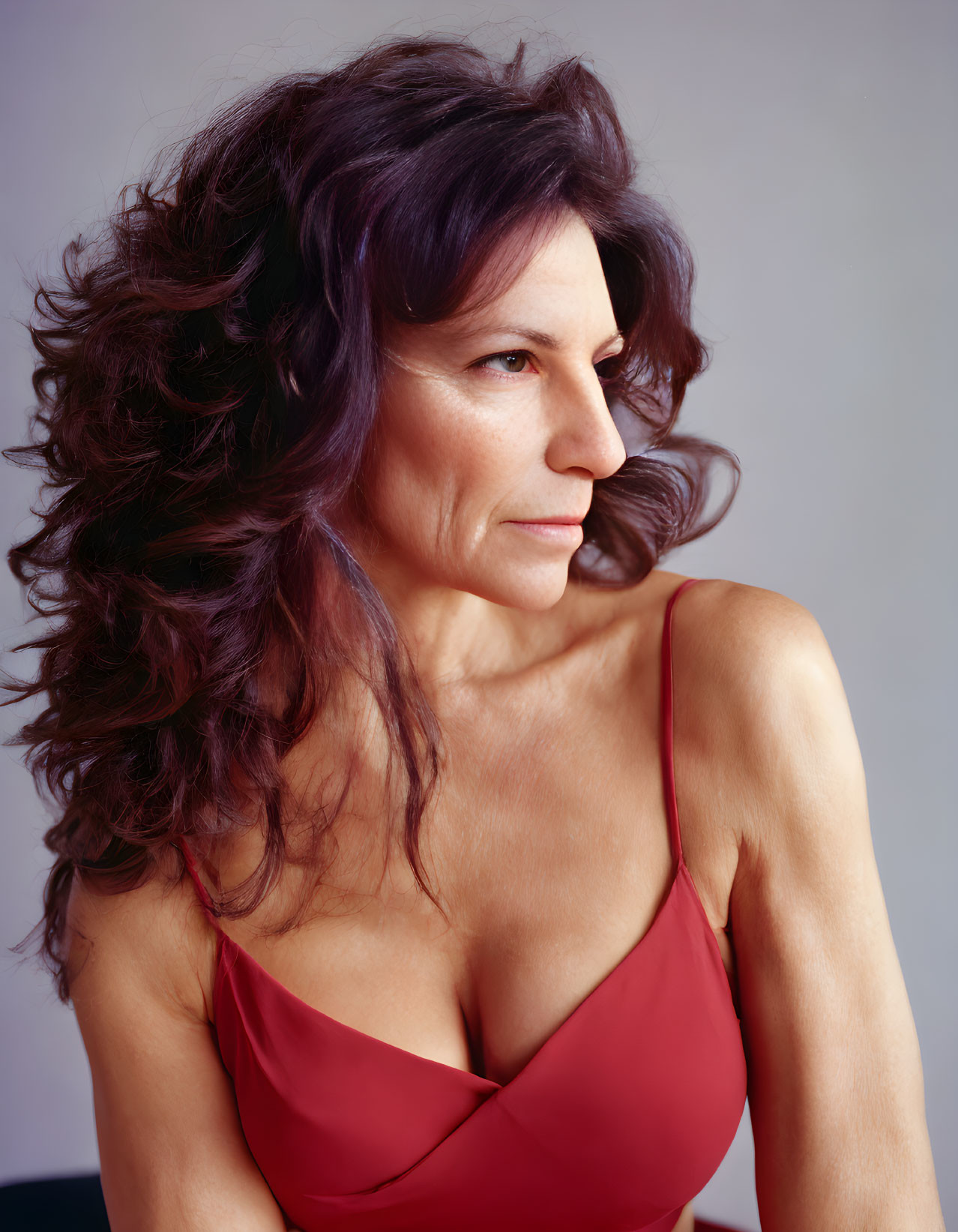 Mature woman with voluminous curly hair in red top on grey backdrop