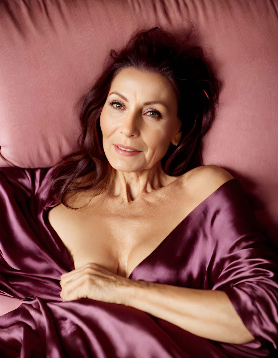 Dark-haired woman lying on pink satin sheet, smiling at camera