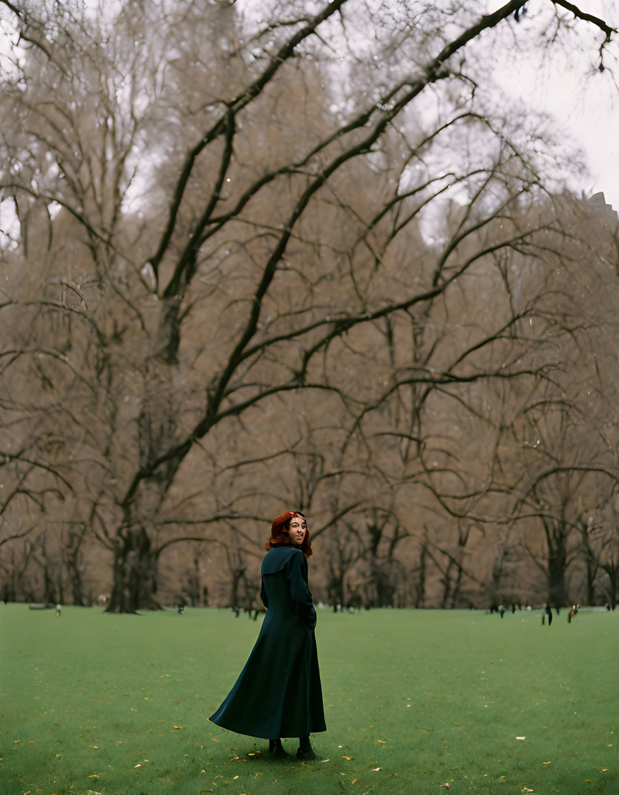 Woman in dark coat standing in park with bare trees and green lawn