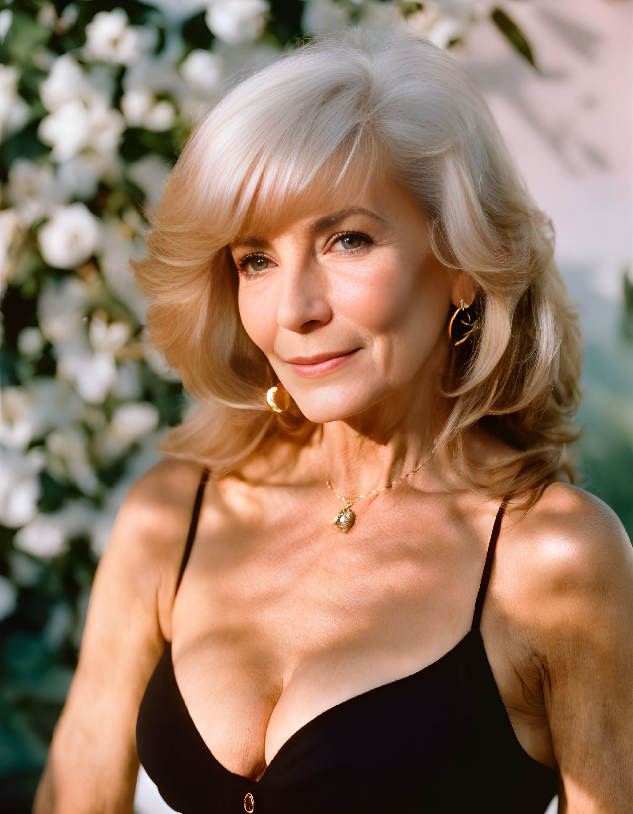 Elegant older woman with blonde hair in black top, hoop earrings, necklace, white flower backdrop