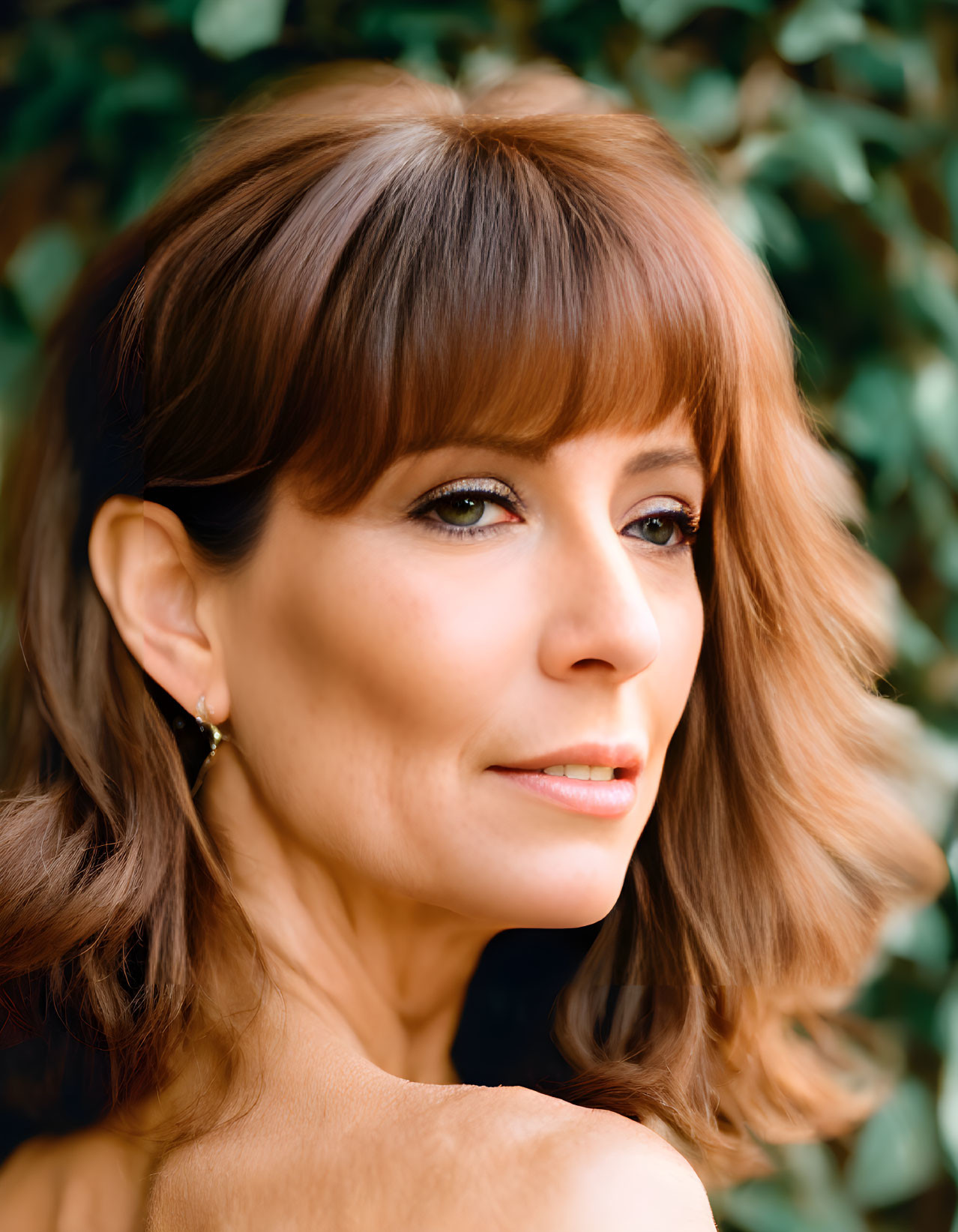 Brown-haired woman with bangs in subtle makeup against green leafy background