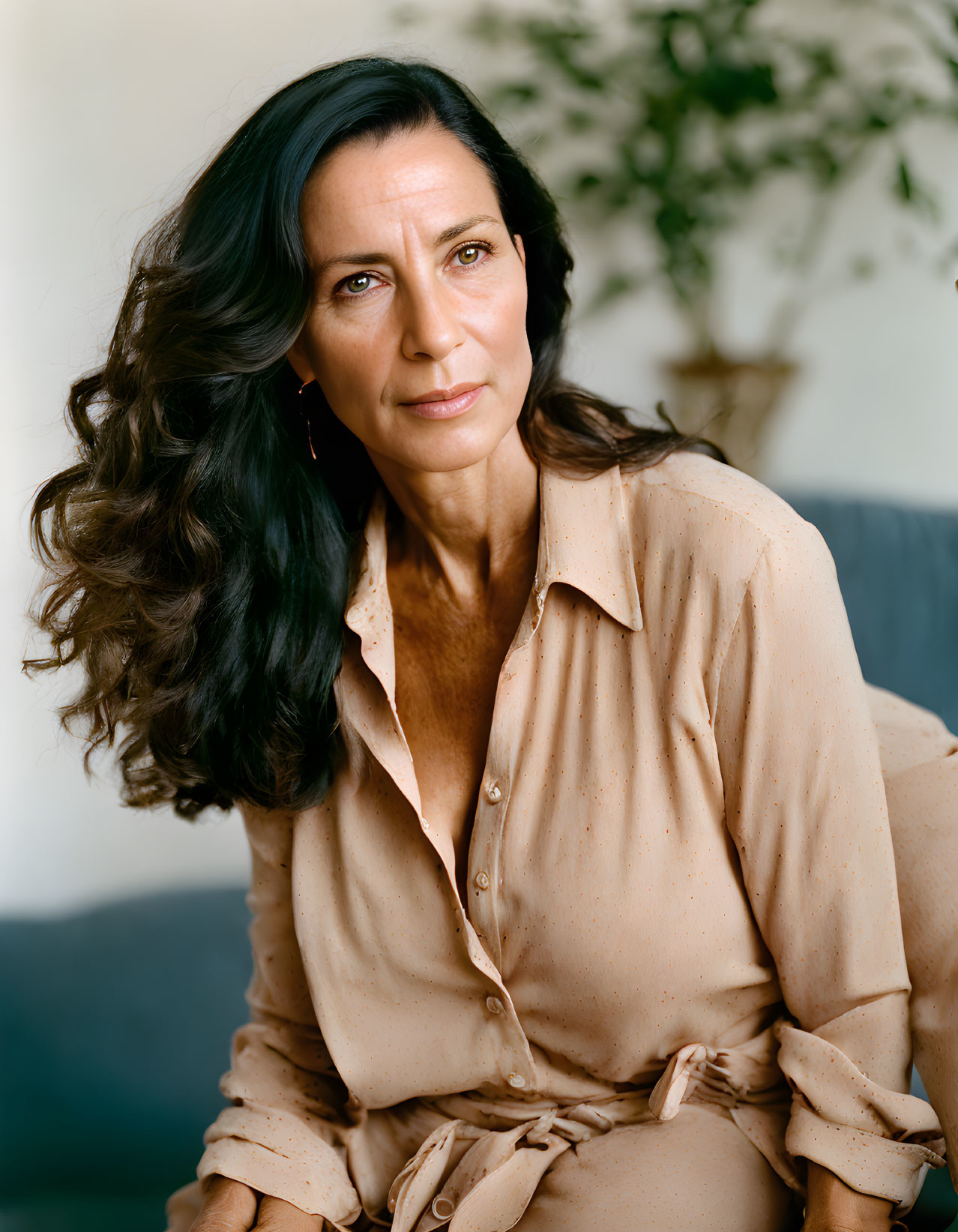 Dark-haired woman in tan blouse against greenery and sofa background