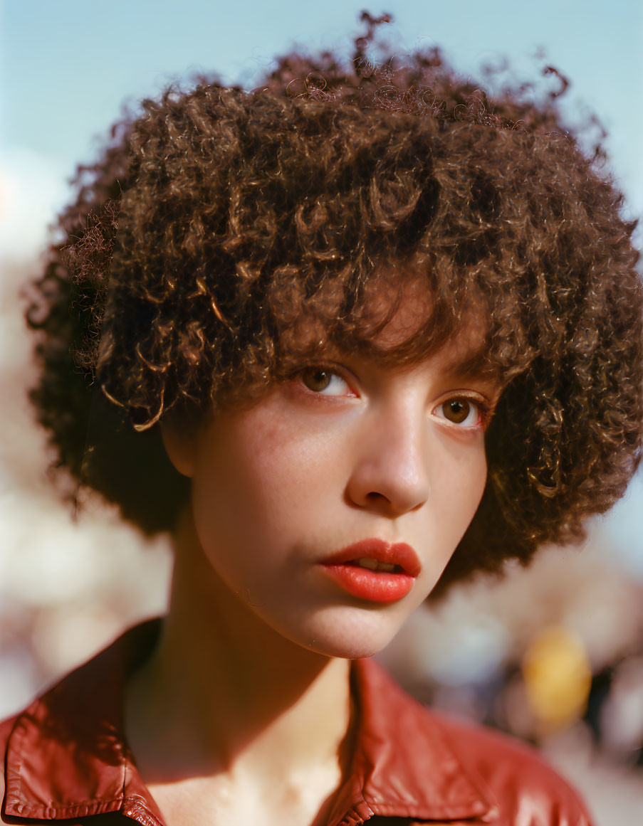 Curly-haired person in red lipstick and jacket gazes at camera