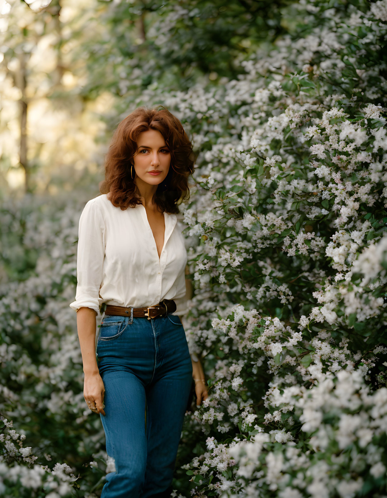 Curly-haired woman in white blouse and jeans among white flowers.