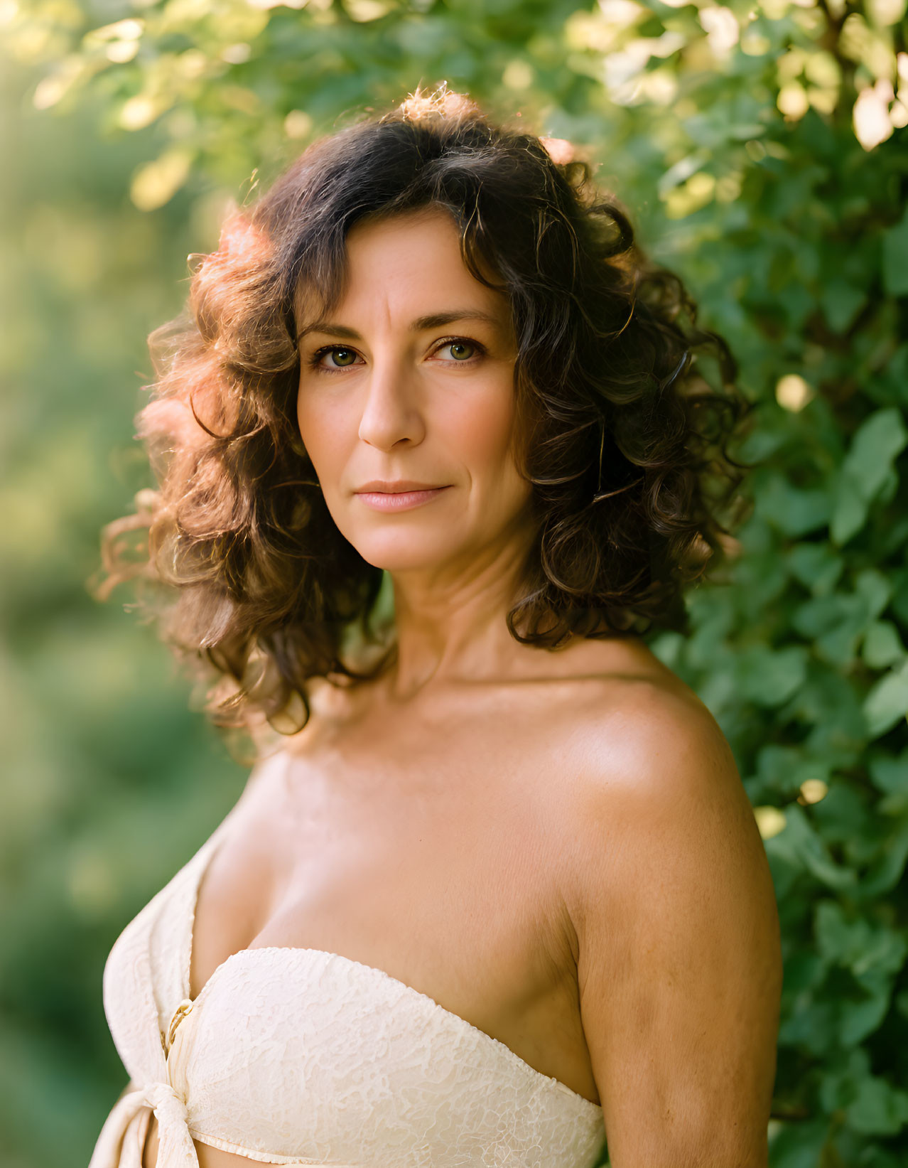 Curly Haired Woman Smiling in Cream Dress on Green Leafy Background