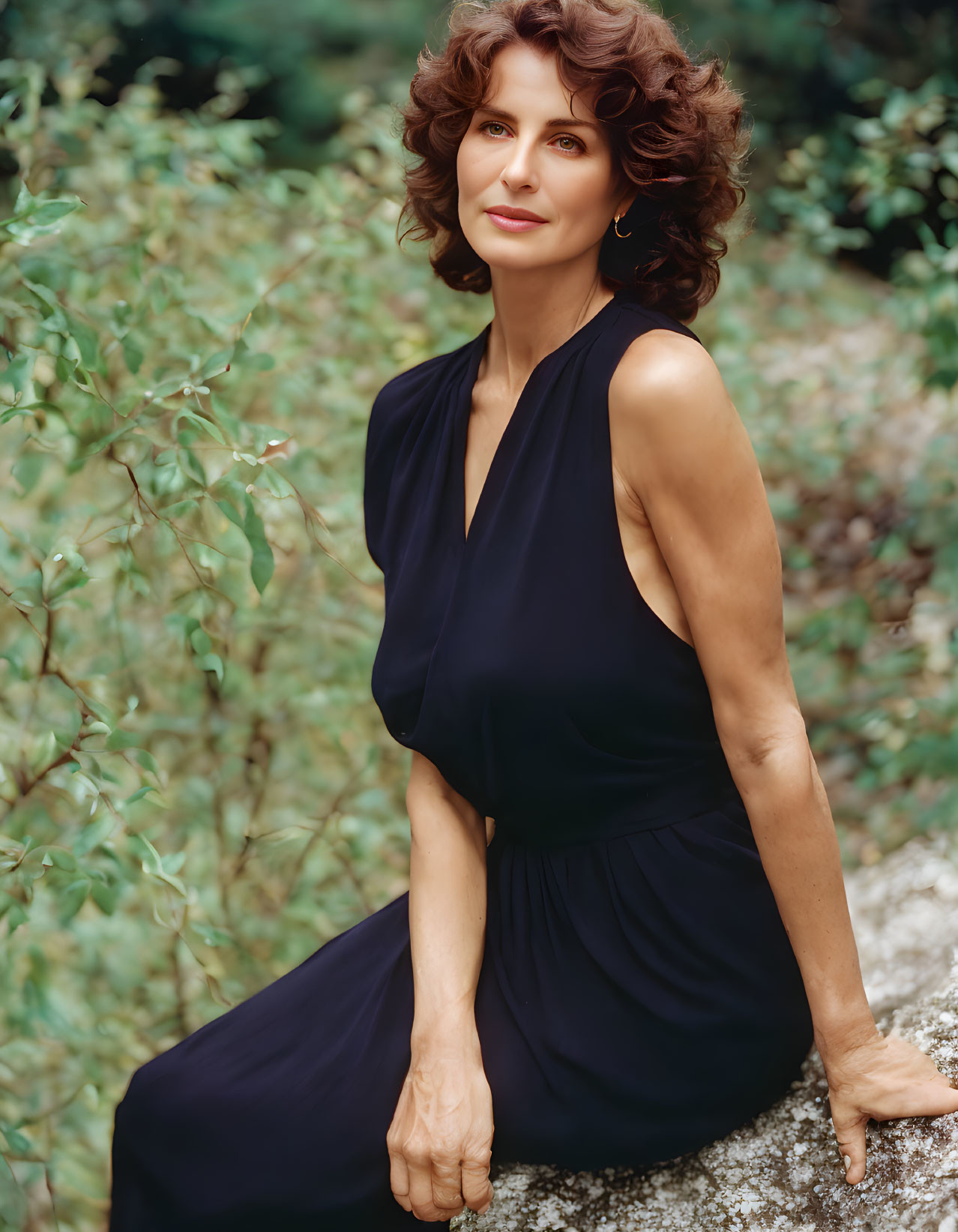 Curly-haired woman in navy dress sitting on rock surrounded by foliage