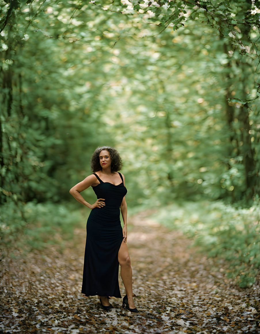 Confident woman in black dress poses in lush green forest with dappled sunlight.