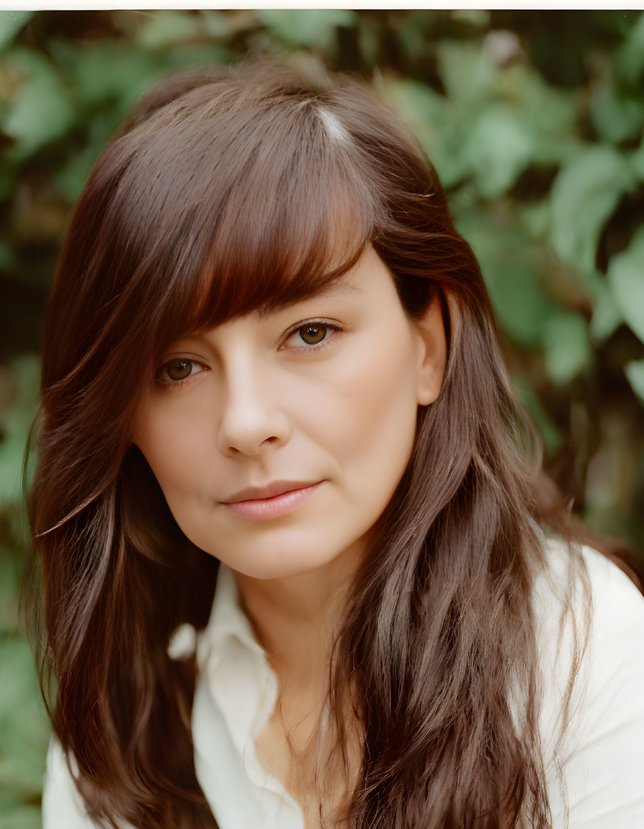 Woman with long brown hair and bangs in light blouse against green leafy background