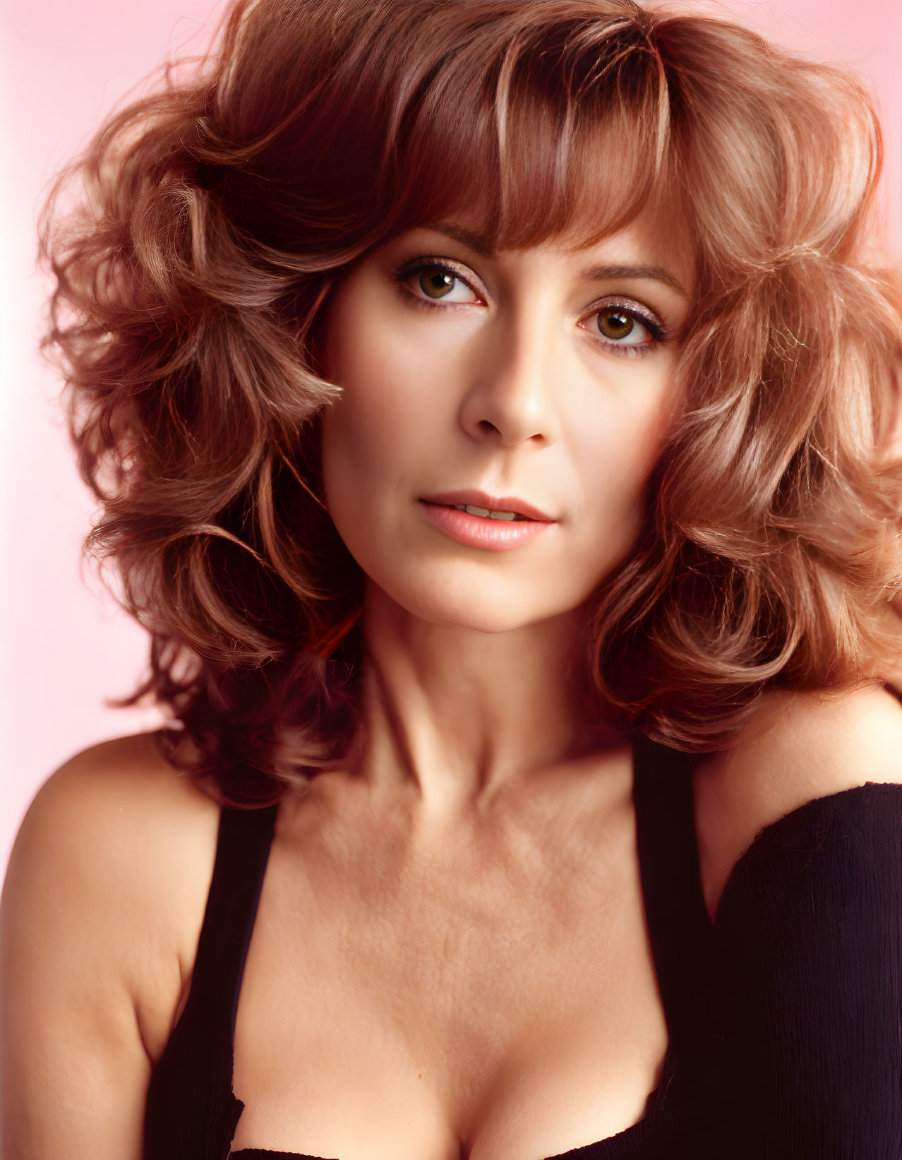 Curly-haired woman in black top on pink background gazes at camera