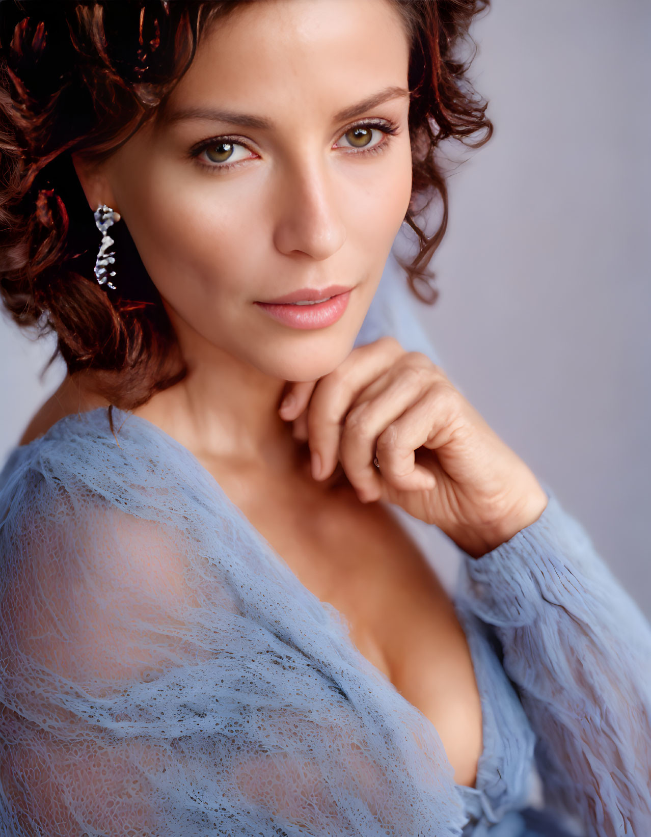 Curly-haired woman in blue lace top with diamond earring posing.