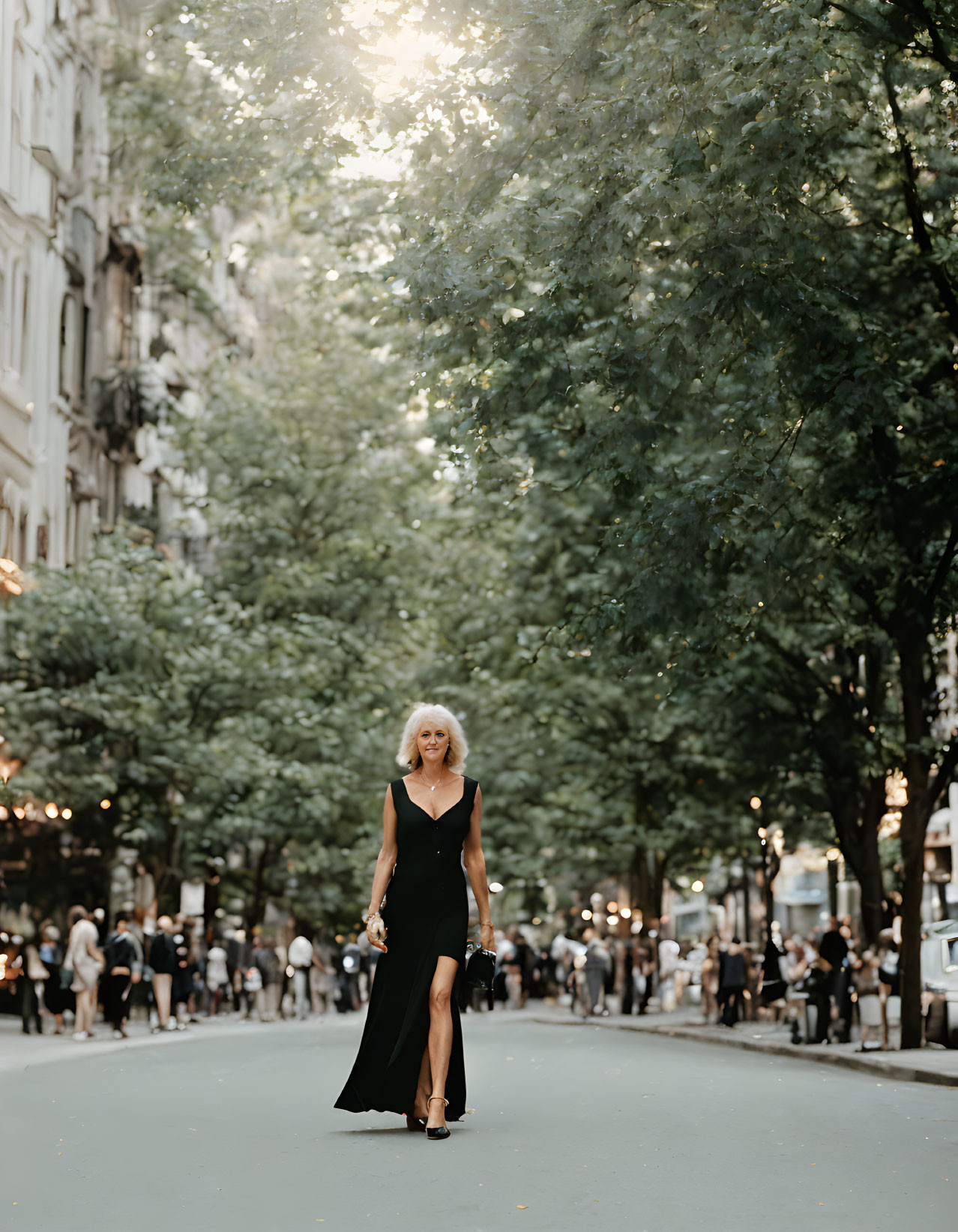 Woman in black dress on tree-lined street with passersby and sunlight.