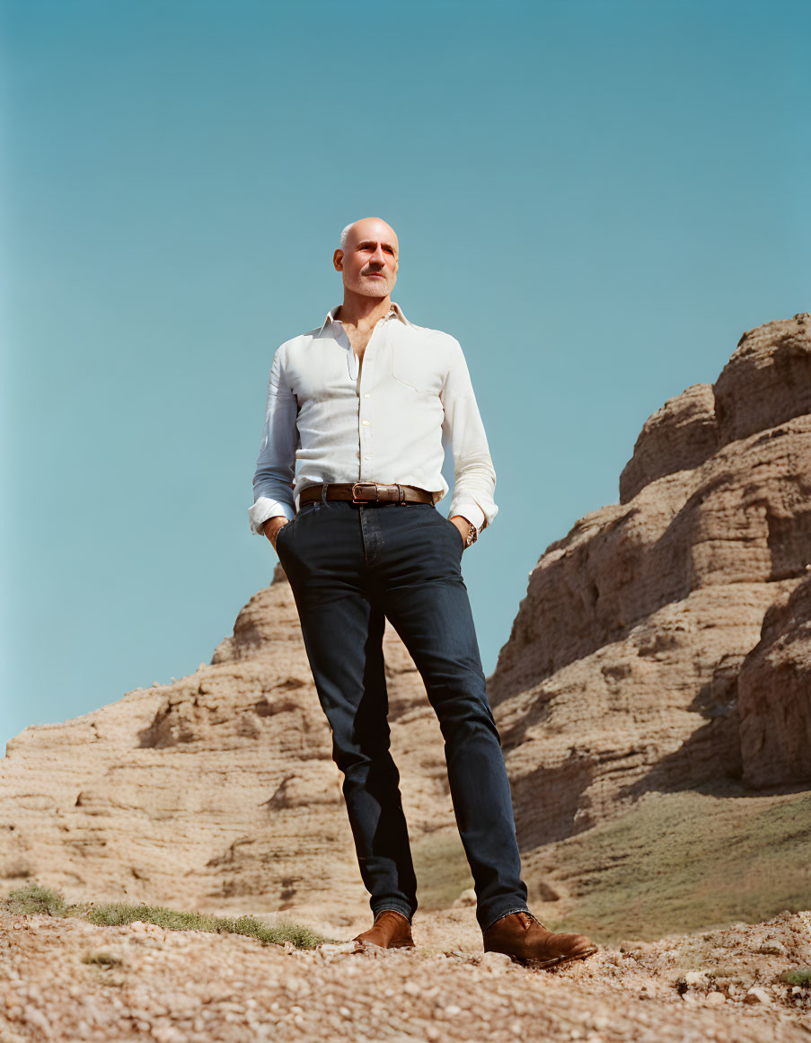 Bald man in white shirt and blue jeans against rocky hills and blue sky