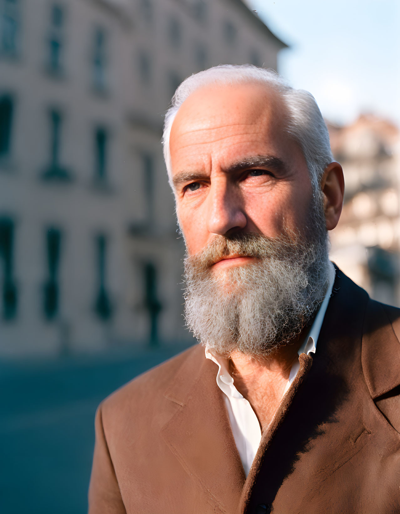 Elderly man in white beard and brown suit outdoors with blurred city background