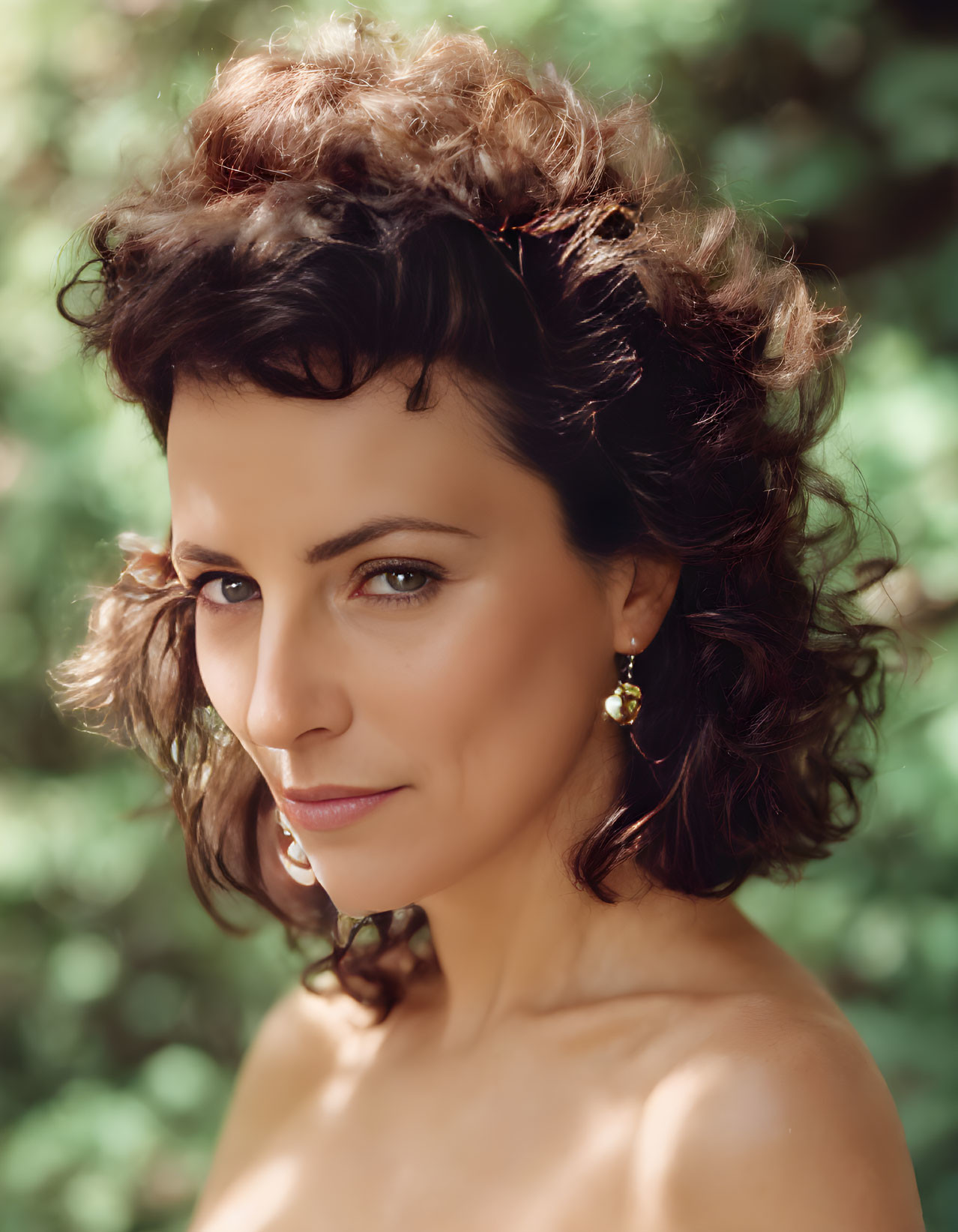 Curly-haired woman portrait with bare shoulders and subtle smile against green foliage.