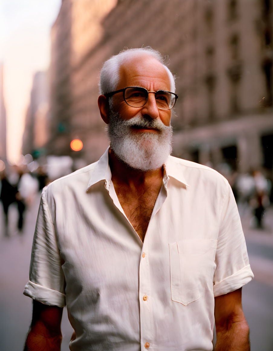 Elderly man with white beard and glasses smiling on city street at dusk