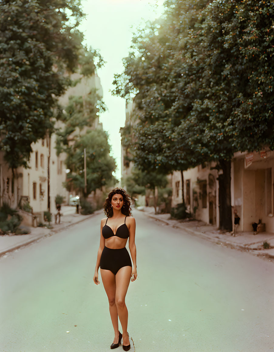 Confident woman in black swimsuit on urban street with trees and sunlight.