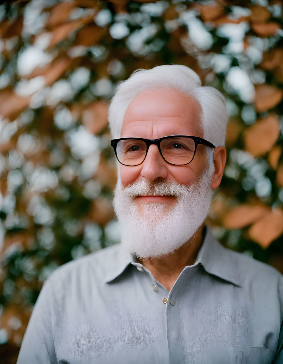 White-bearded elderly man in glasses, smiling in blue shirt against foliage.