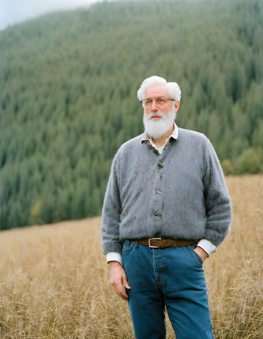 White-bearded elderly man in beret in field with forested hill.