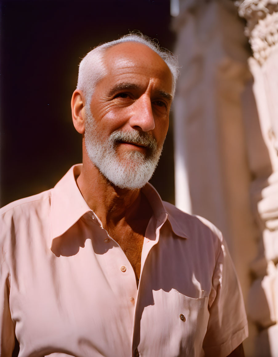 Elderly man with white beard in pink shirt by columned structure