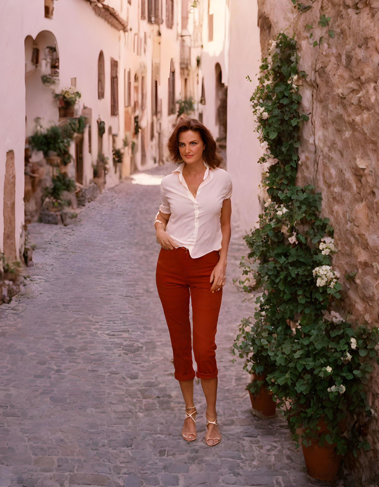 Woman in white blouse and red pants on cobblestone street with old buildings.