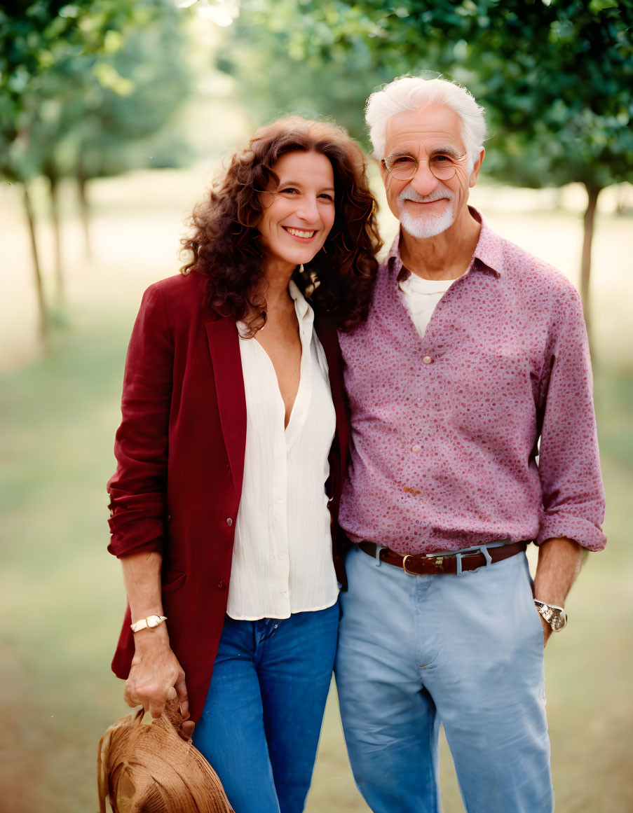 Smiling couple in sunlit orchard: woman with curly hair, red blazer, man with