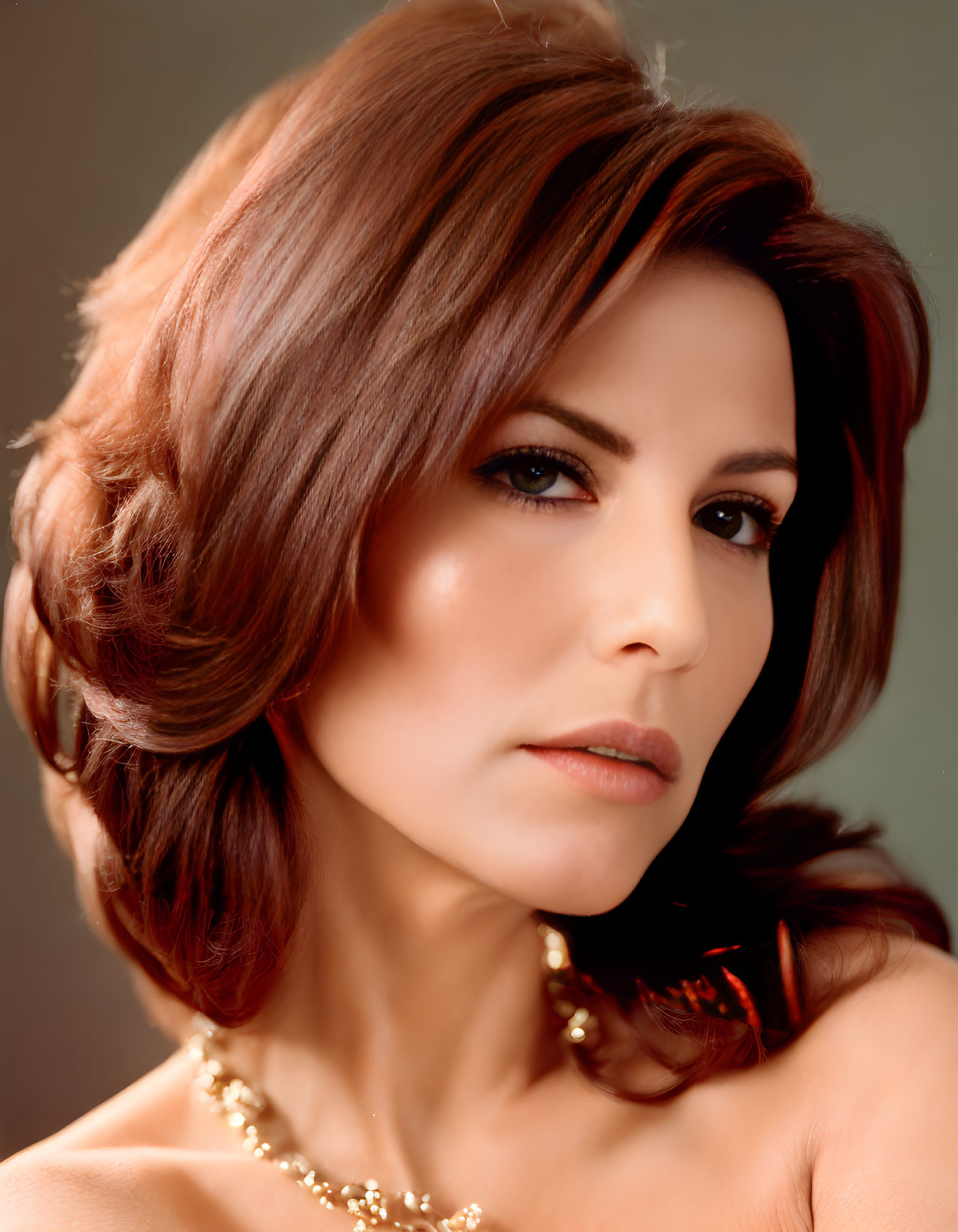 Portrait of woman with auburn hair, gold necklace, and soft gaze on neutral background