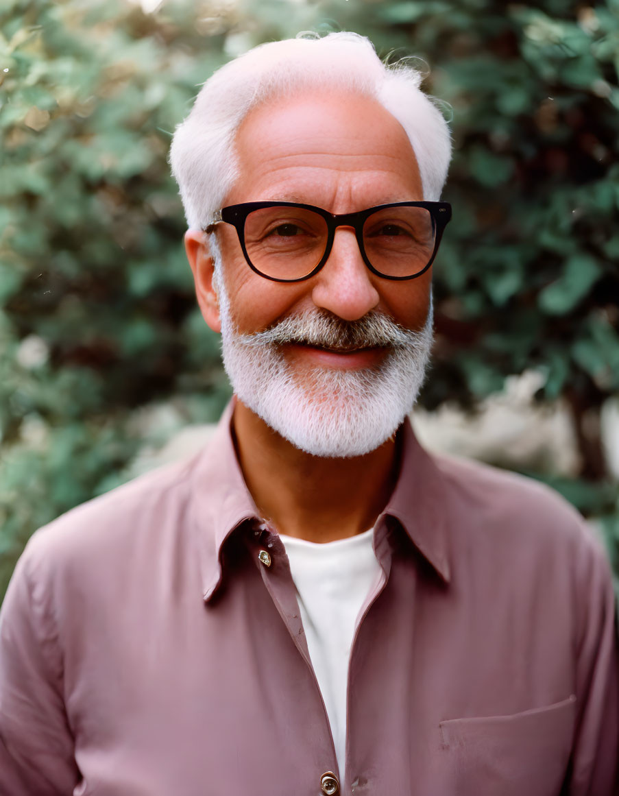 Elderly man with white hair and beard in mauve shirt against green foliage