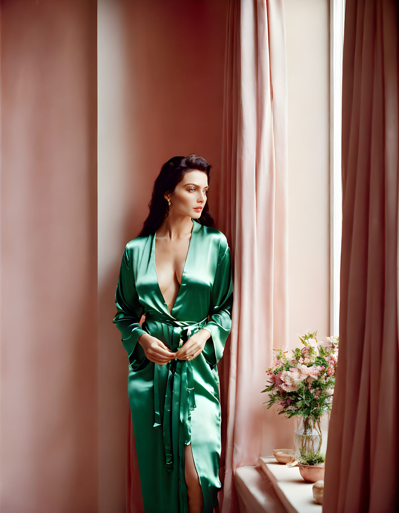 Elegant woman in green dress by window with pink drapes and flowers.