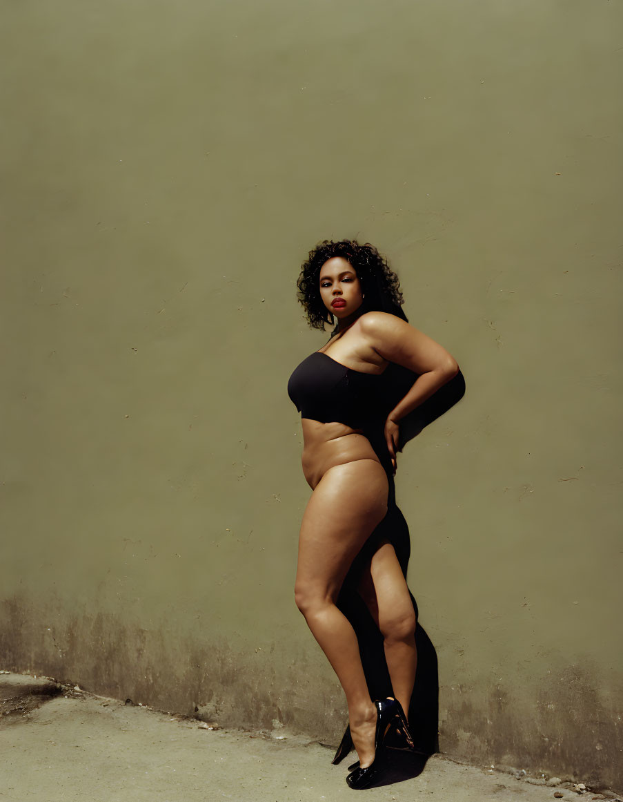 Confident woman in black outfit against beige wall with curly hair and high heels