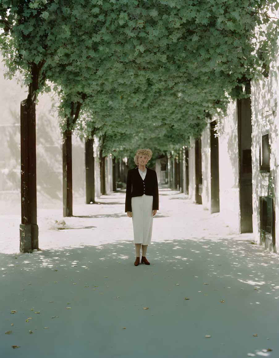 Woman in black top, white pants, and red shoes standing on tree-lined path