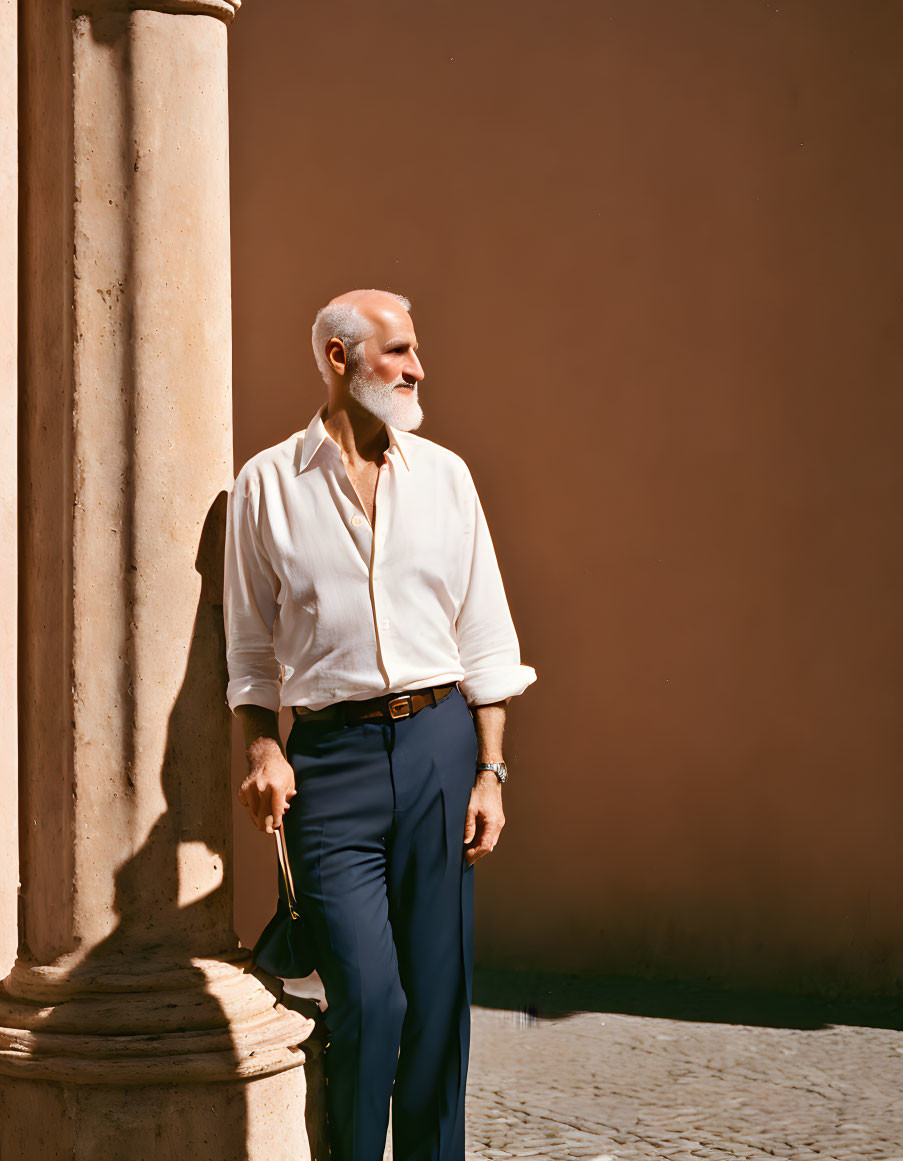 Elderly man with beard leaning against column in white shirt and blue trousers under sunshine.