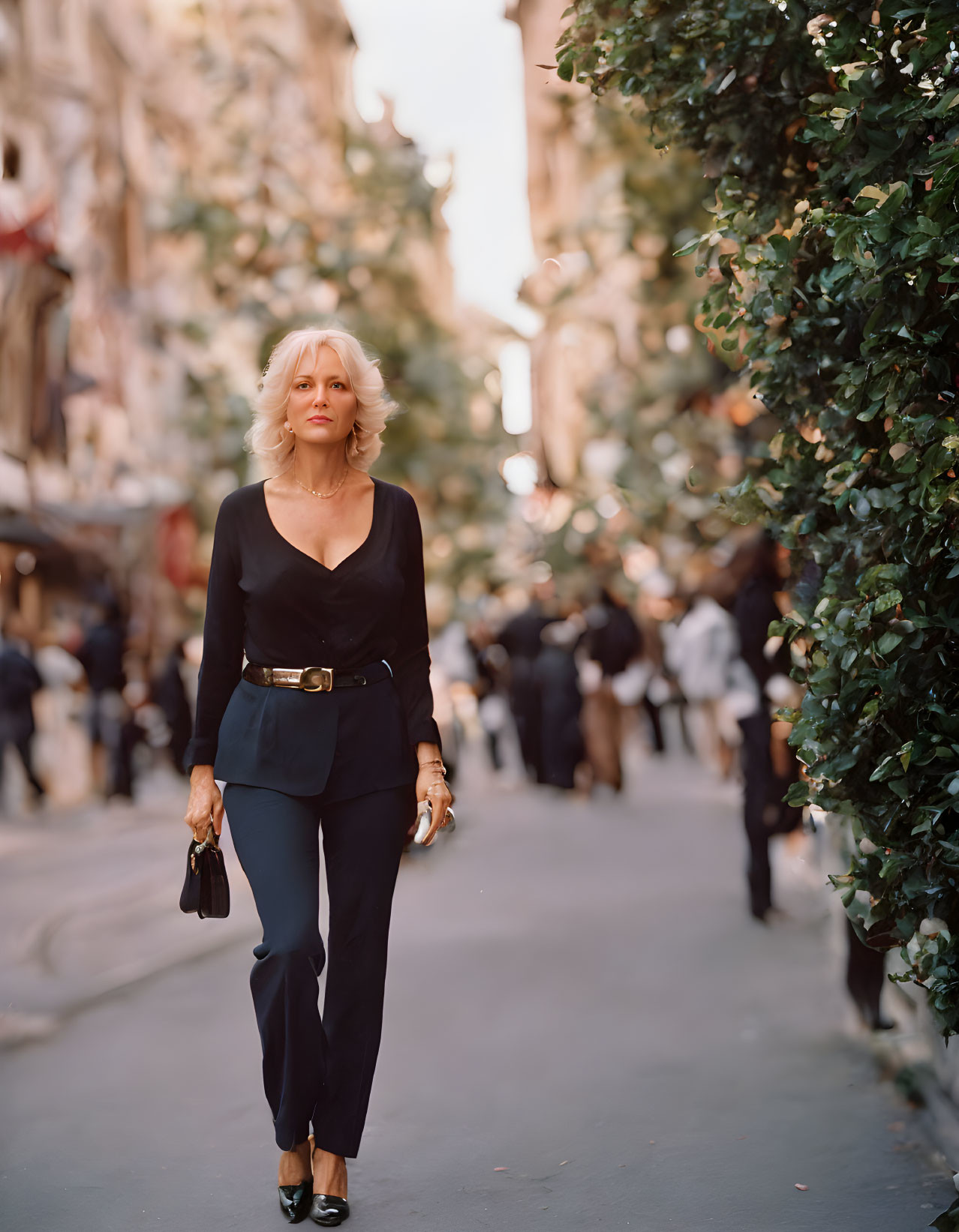 Blonde woman in black top and blue trousers walking on busy tree-lined street