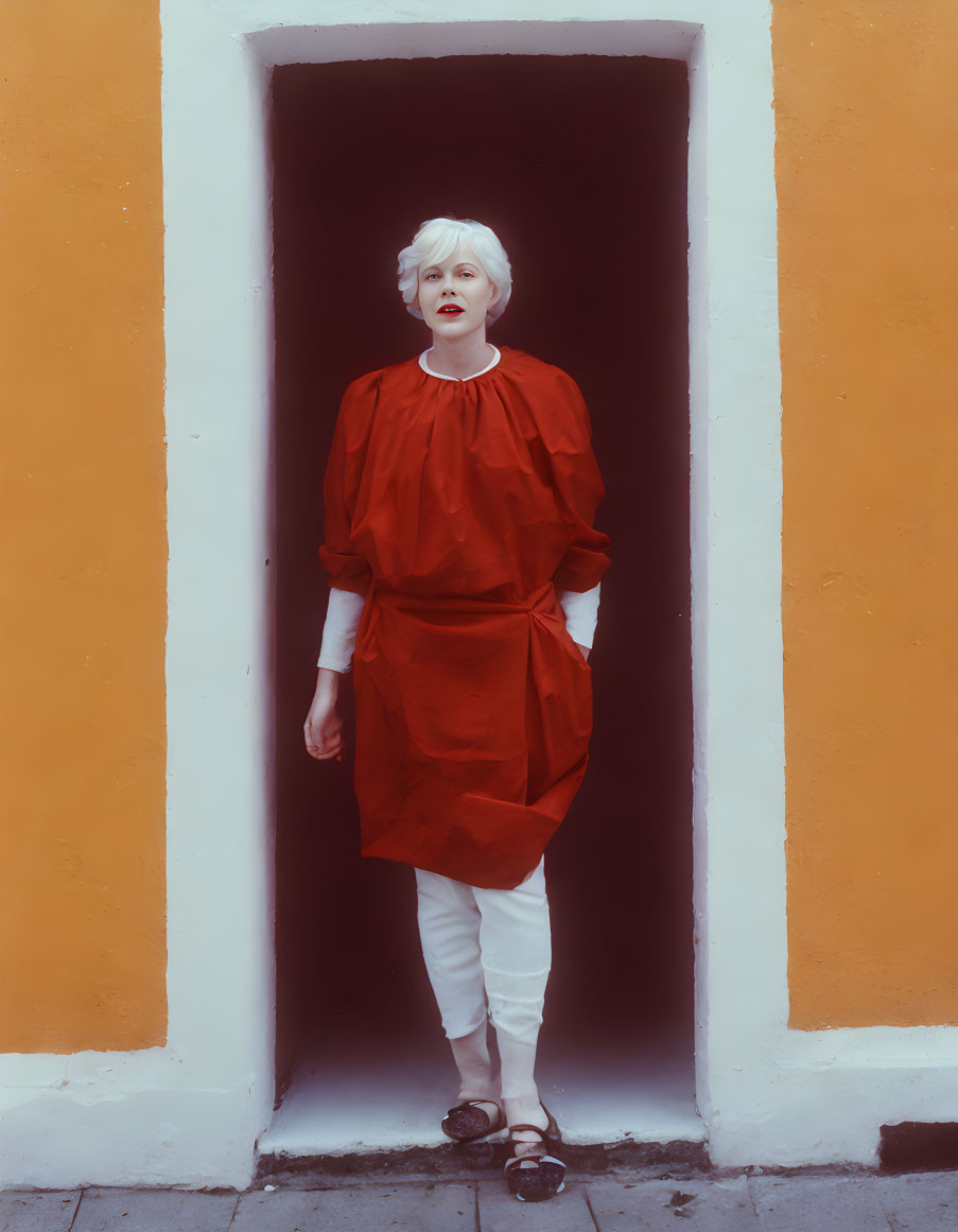 Blonde person in red tunic standing in white-framed doorway