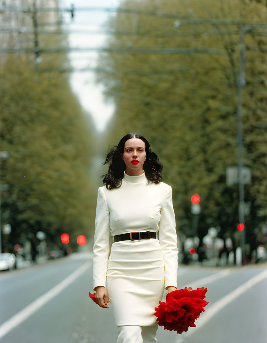 Woman in white dress walking on street with red flowers, leafy trees, and traffic lights.