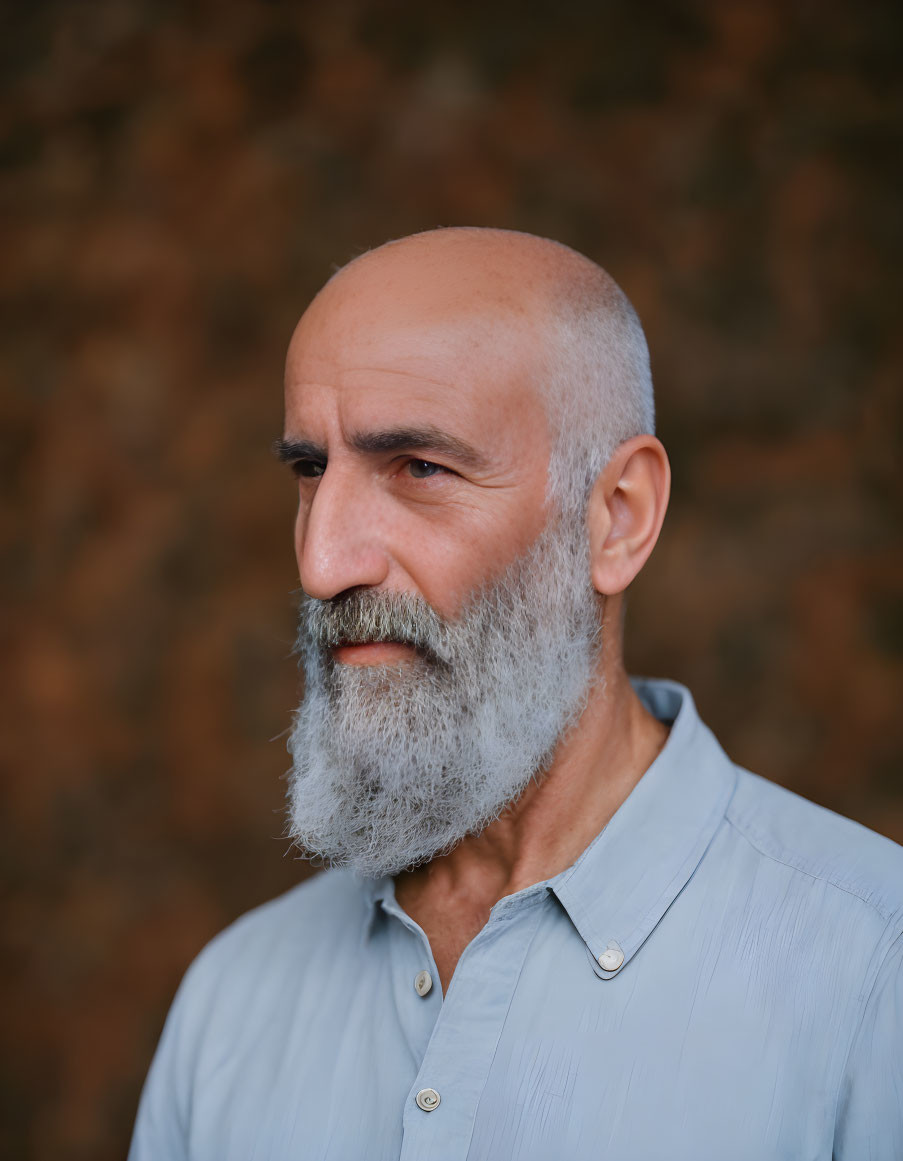 Gray-bearded bald man in light blue shirt on blurred brown background