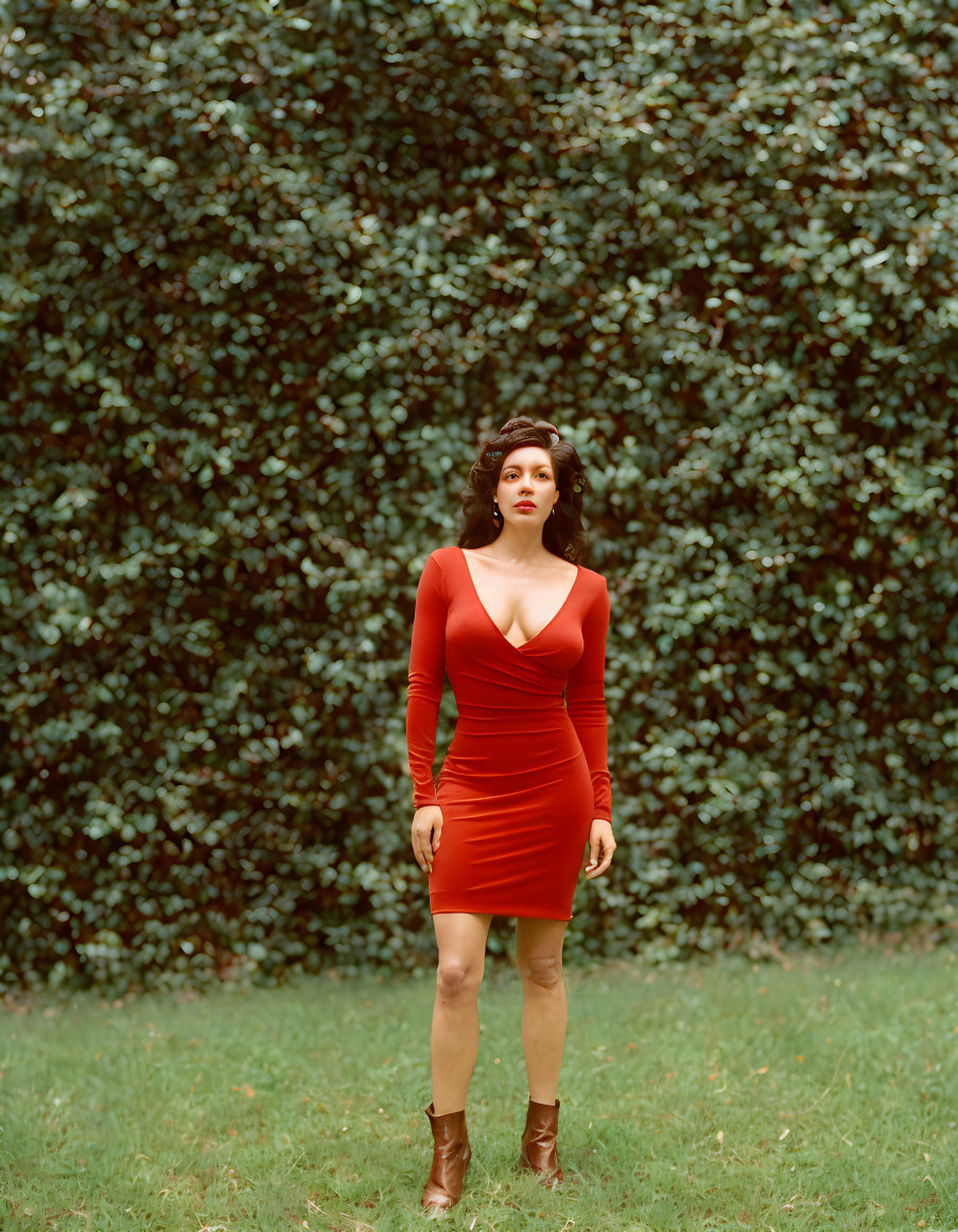 Woman in red dress and brown boots by lush green hedge with wavy dark hair